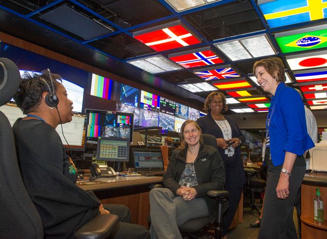 Four women in a modern mission control room with various screens and international flags on the ceiling. Ideal for content about teamwork, STEM, space exploration, and workplace diversity. Highlights collaboration and the integration of diverse skills in high-tech environments.