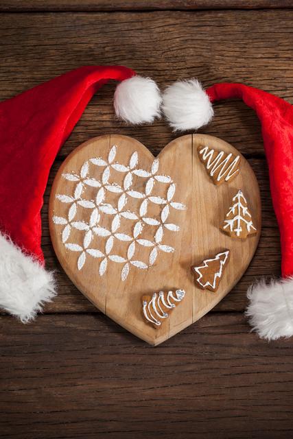 Heart-shaped wooden board with gingerbread cookies decorated with icing, flanked by two Santa hats. Ideal for holiday-themed promotions, Christmas cards, festive blog posts, or seasonal marketing materials.