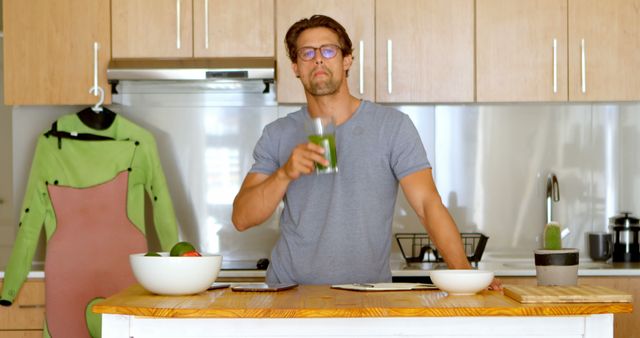 Man drinking a green smoothie in a modern kitchen setting. He wears glasses and a casual grey shirt, indicating a health-conscious lifestyle. Suitable for health and wellness promotions, nutrition blogs, and advertisements for kitchen appliances or fitness-related products.