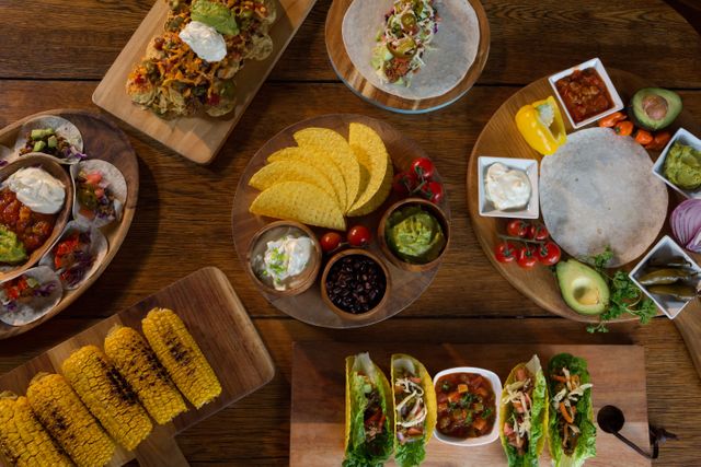 Assorted Mexican dishes including tacos, nachos, guacamole, salsa, and grilled corn arranged on a wooden table. Ideal for use in food blogs, restaurant menus, culinary magazines, and festive event promotions.