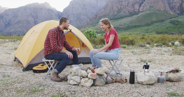 Young couple sitting by campfire outside tent in mountainous area, enjoying morning conversation and nature. Perfect for promoting camping gear, outdoor adventures, eco-tourism, or showcasing relaxation and lifestyle.