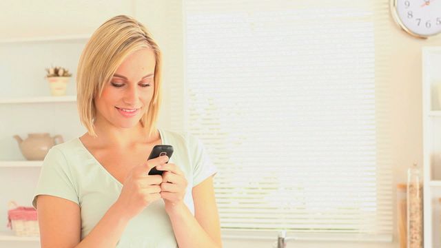 Fairhaired woman smiling while sending text messages in a bright, modern kitchen. Ideal for depicting everyday use of technology, communication, and personal time at home. Great for advertising mobile apps, home lifestyles, and technology usage.