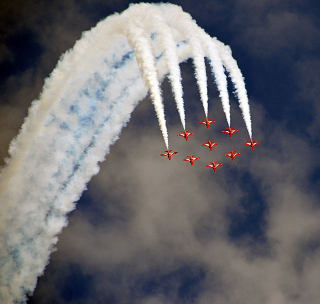 A fleet of red jets engaged in aerobatic performance with trailing smoke, creating an arc pattern against the backdrop of a partly cloudy sky. Ideal for use in articles about airshows, aviation-themed events, teamwork and coordination in flight, or promoting air festivals and aviation exhibitions.