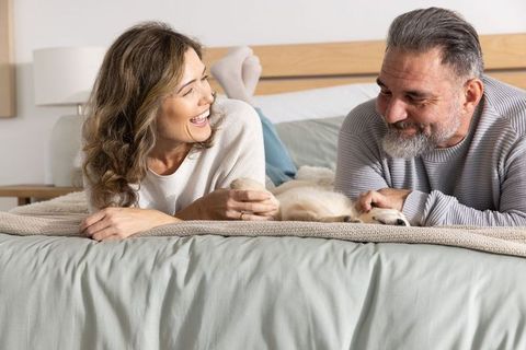 Middle-aged couple laughing petting puppy on cozy bed, intimate morning moment