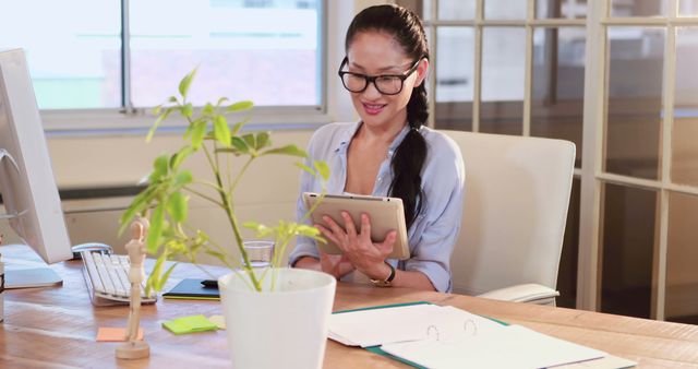 Young professional woman using tablet at modern office desk. She is smiling and appears engaged with her work, indicating productivity and modern work environment. Suitable for themes related to business, technology, professional life, remote work, success, and career development. The presence of the potted plant adds a touch of natural element and aesthetic to the workspace.