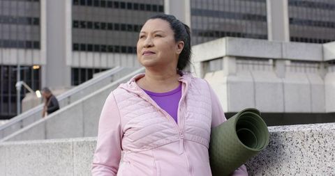 Asian Senior Woman Carrying Yoga Mat in Urban Plaza Wearing Pink Quilted Jacket