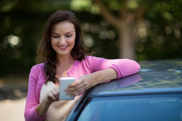 Woman standing by car, smiling while using mobile phone. Ideal for themes related to communication, technology, lifestyle, and outdoor activities. Suitable for advertisements, blogs, and articles focusing on mobile technology, connectivity, and casual lifestyle.