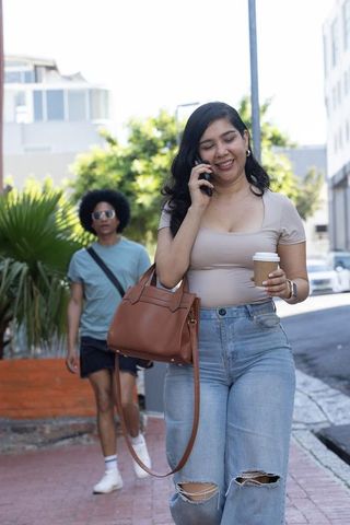 Young woman walking and talking on phone holding coffee and handbag in urban street