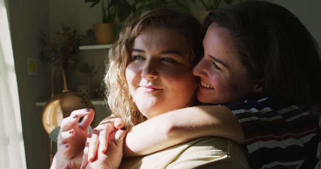 Image shows a cheerful lesbian couple embracing in a warmly lit living room. The bright light enhances their happy expressions and strong bond. Ideal for use in articles, blogs, or advertisements focusing on relationships, LGBTQ+ rights, same-sex couple life, and emotional connections.