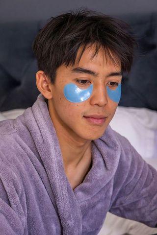 Young asian man using blue hydrogel under-eye patches in lavender bathrobe