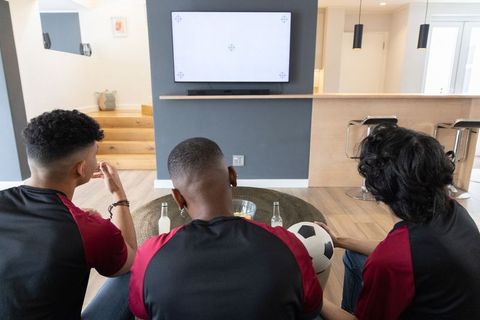 Diverse friends watching soccer on wall-mounted tv wearing matching jerseys in living room