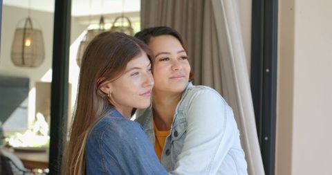 Female couple embracing by glass door in cozy open-plan home wearing denim jackets