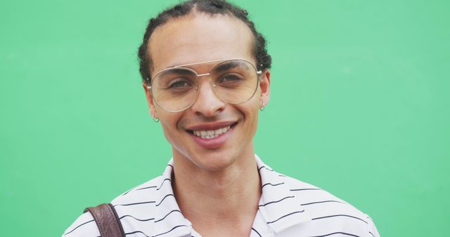 Smiling man with glasses and curly hair standing in front of green background. Wearing striped shirt and carrying a bag over shoulder. Perfect for design projects, advertising campaigns, fashion magazines, and lifestyle blogs.