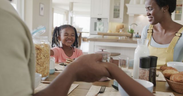 Happy African American father, mother, and daughter are having breakfast together at home, sharing a joyful moment. Parents holding hands, little girl in the middle of laughter and conversation at the table. Ideal for depicting family life, happiness and togetherness. Perfect visuals for articles on family bonds, morning routines, healthy living, and multicultural representation.