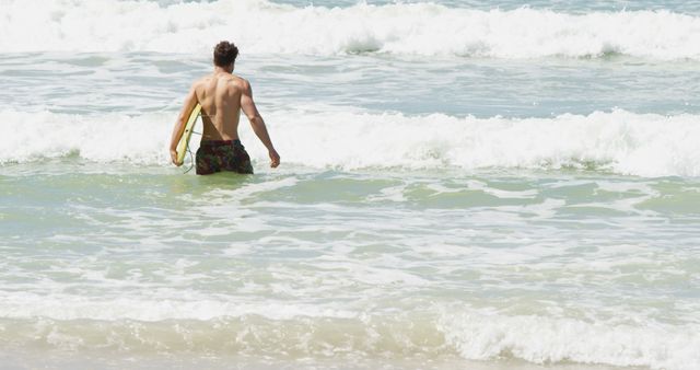 A young Caucasian male is seen entering the ocean waves with a surfboard, with copy space. His relaxed posture and the clear day suggest an enjoyable surfing session ahead.