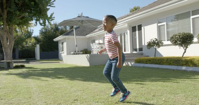 Child wearing striped shirt smiling and jumping joyfully on well-maintained lawn outside modern home on sunny day. Great for advertisements related to children's activities, outdoor fun, family life, summer vacations, and real estate.