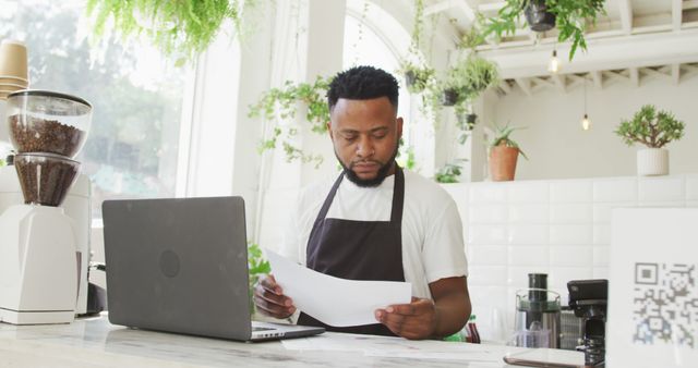 African american male cafe owner using laptop and looking at paperwork behind counter at cafe. small independent cafe business.