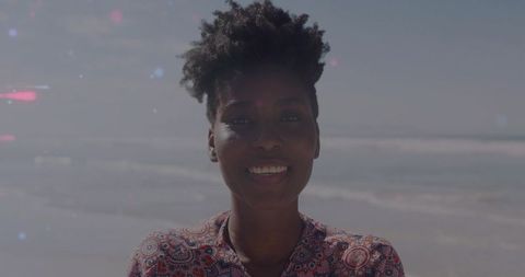 Smiling woman standing at shoreline wearing patterned blouse with ocean horizon and magenta flare