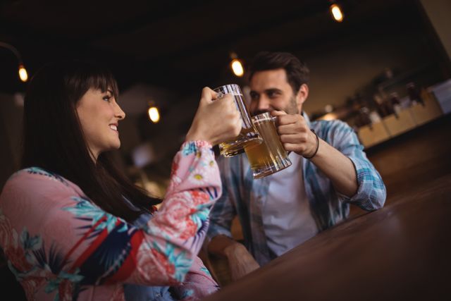 Couple enjoying a night out at a bar, toasting with glasses of beer. Ideal for use in advertisements for bars, pubs, or breweries, as well as articles or blogs about socializing, relationships, and nightlife.