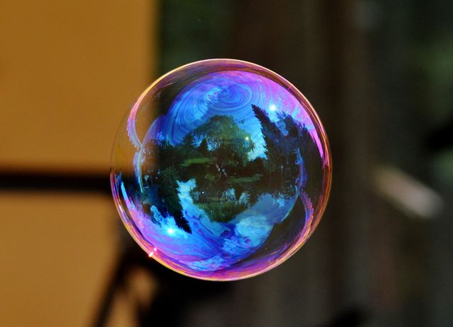 Up-close perspective of a single soap bubble reflecting vibrant colors and the serene outdoor environment of trees and nature. Useful for themes related to relaxation, childhood, environmental awareness, and beauty in simplicity. Perfect for backgrounds, posters, and educational materials.