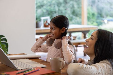 Indian mother and daughter enjoying online learning, child putting on headphones by laptop