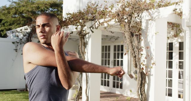 Young man in a tank top stretching outdoors near a white house with vines. This is an ideal image for topics related to fitness, healthy living, outdoor activities, exercise routines, and warm-up exercises. Perfect for fitness blogs, workout routines, health and wellness articles, and promoting a healthy lifestyle.