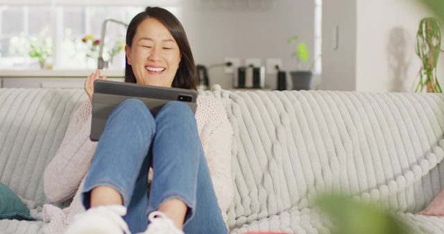 This image shows a young woman sitting comfortably on a couch while using a tablet at home. She appears relaxed and happy, making it ideal for use in lifestyle blogs, technology advertisements, home decor magazines, and any content emphasizing modern living or casual relaxation. Perfect for illustrating digital connectivity in everyday life.