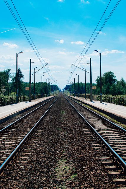 Image showcases a symmetrical perspective view of empty railroad tracks extending towards the horizon under a clear blue sky. This setting evokes feelings of travel, adventure, and solitude, suitable for use in travel promotions, transportation industry content, motivational posters, or artwork emphasizing journeys and paths.
