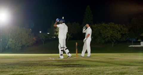 Diverse teammates adjusting helmet at crease under floodlights during night cricket match