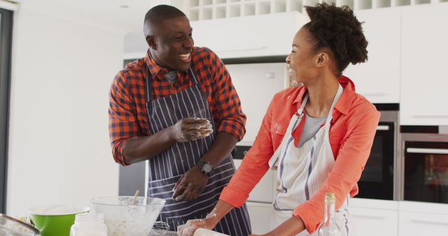 This lively image depicts an African American couple joyfully baking together in their kitchen. They seem to be engrossed in a fun moment, playing with flour and laughing heartily. This engaging scene can be used in promotional materials for relationship blogs, cooking websites, advertisements for kitchen appliances, or articles focusing on family bonding and quality time spent at home. It highlights themes of love, happiness, and the joys of everyday life.