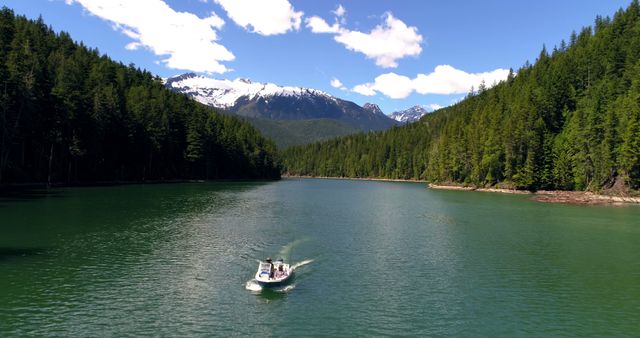 Boat cruising on calm waters of a lake surrounded by dense forest with snow-capped mountains in background. Ideal for use in travel brochures, nature blogs, and promotional materials for outdoor activities. Perfect for showcasing serene landscapes, adventure, and recreational boating.