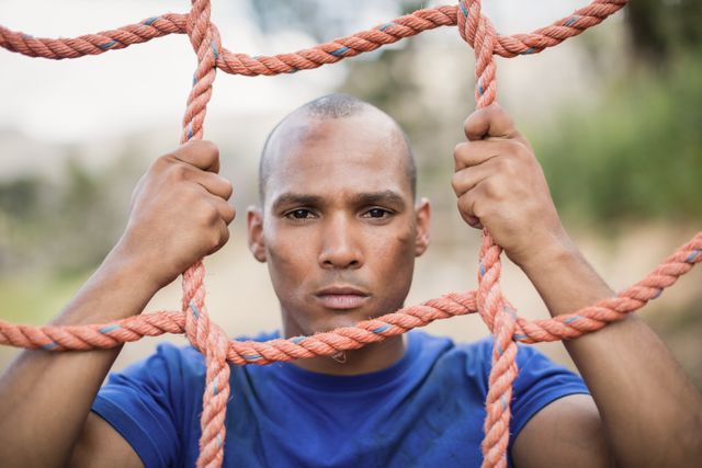 This image depicts a fit man climbing a net during an obstacle course in a boot camp. His expression shows determination and focus, making it ideal for use in fitness and training-related content. It can be used in advertisements for boot camps, fitness programs, or motivational materials promoting physical activity and perseverance.