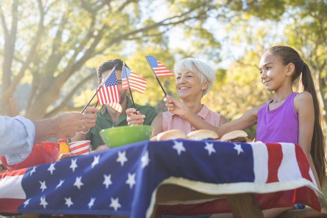 Family enjoying a picnic in the park, holding American flags and smiling. Ideal for themes of patriotism, family bonding, summer activities, and holiday celebrations. Perfect for use in advertisements, social media posts, and articles related to American holidays, family gatherings, and outdoor events.