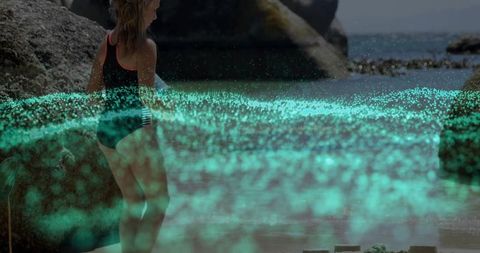 Girl standing on rocky beach watching glowing bioluminescent water and turquoise sparkles