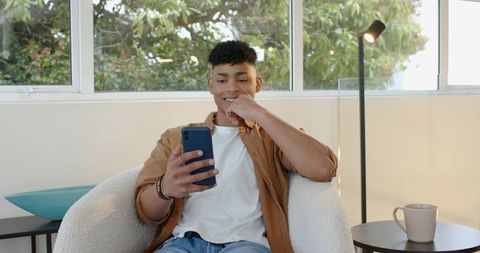 Young man relaxing in modern sunlit living room scrolling smartphone and smiling