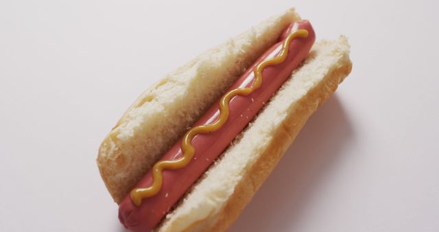 Depicts a hot dog with mustard on top, placed in a soft bun against a white background. Ideal for advertisements related to fast food, culinary articles, and promotions for food-related businesses. Can be used in menus, flyers, and social media posts to showcase a simple, classic American meal.