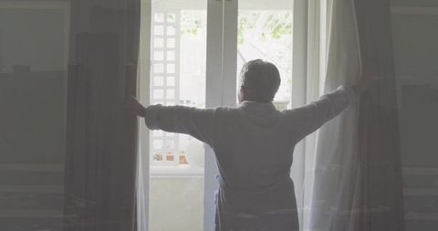 Senior woman opening curtains at balcony door in robe morning light silhouette