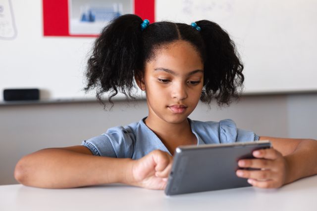 Biracial elementary schoolgirl using digital tablet while studying at desk in classroom. unaltered, education, learning, studying, concentration, wireless technology and school concept.