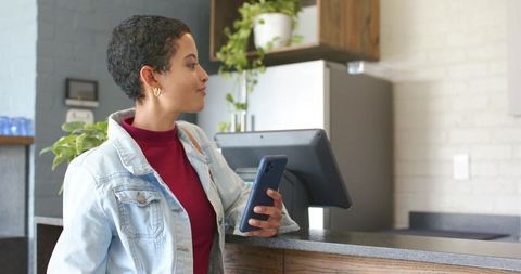Young woman making mobile payment at cafe counter with smartphone and touchscreen pos