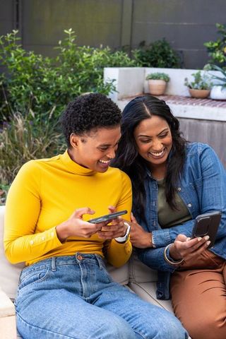 Diverse female friends sharing smartphones and laughing on outdoor patio bench