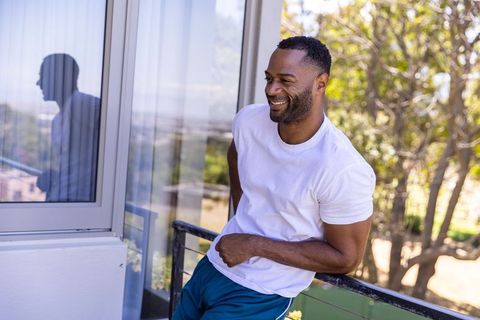 African man leaning on balcony railing smiling, enjoying sunny terrace with greenery