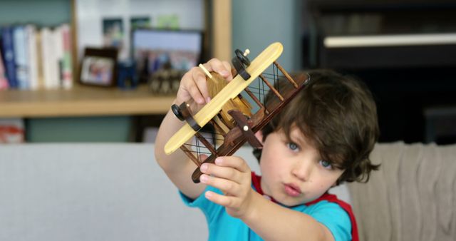 Young boy enthusiastically playing with a wooden toy airplane while sitting on a couch in a living room. Ideal for use in educational materials, parenting blogs, children's product advertisements, or articles about creativity and imaginative play in children.