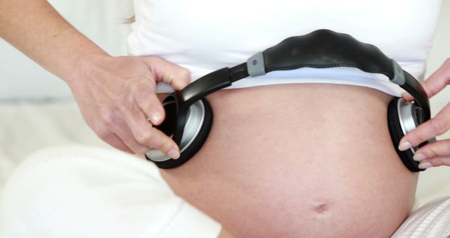 Pregnant woman holds headphones on her belly, possibly playing music or soothing sounds for the baby. Useful for articles, blog posts, or educational materials on prenatal care, music therapy, or maternity wellbeing.