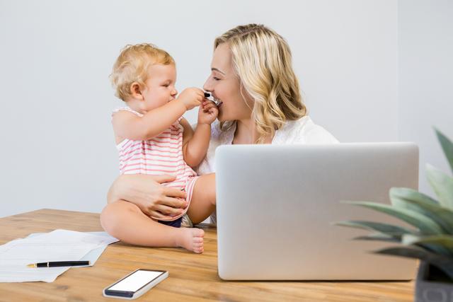 Mother holding baby girl while working on laptop at home. Ideal for articles on work-life balance, remote work, parenting tips, family bonding, and modern motherhood. Can be used in blogs, social media posts, and advertisements promoting family-friendly work environments.