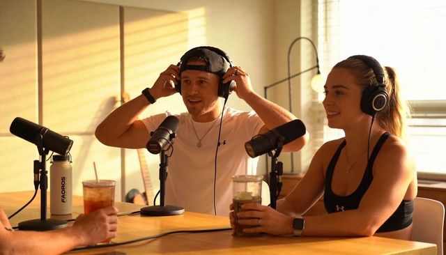 Cohosts recording podcast adjusting headphones in sunlit home studio with casual drinks