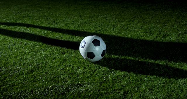 A classic black and white soccer ball rests on a lush green field illuminated by a dramatic spotlight, creating a long shadow. The image captures the anticipation and excitement associated with soccer matches played under the lights.