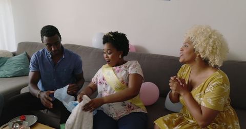 Black friends celebrating baby shower at home unwrapping gift, honoree wearing yellow sash