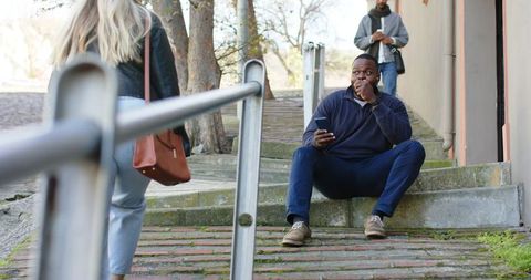 African american man sitting on brick steps checking smartphone as passerby walks past