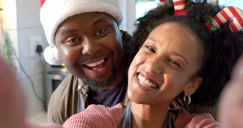 Joyful diverse couple taking festive selfie in cozy kitchen wearing Santa hat and apron