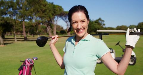 A woman, smiling brightly, stands confidently on a lush, green golf course. She casually rests a golf club on her shoulder, exuding relaxation and enjoyment. With a backdrop of vibrant trees and a golf cart, this scene captures the serene beauty and leisurely pace of golfing. Ideal for promotional materials in sports, retirement communities or leisure activities, it illustrates joy and relaxation associated with golf.